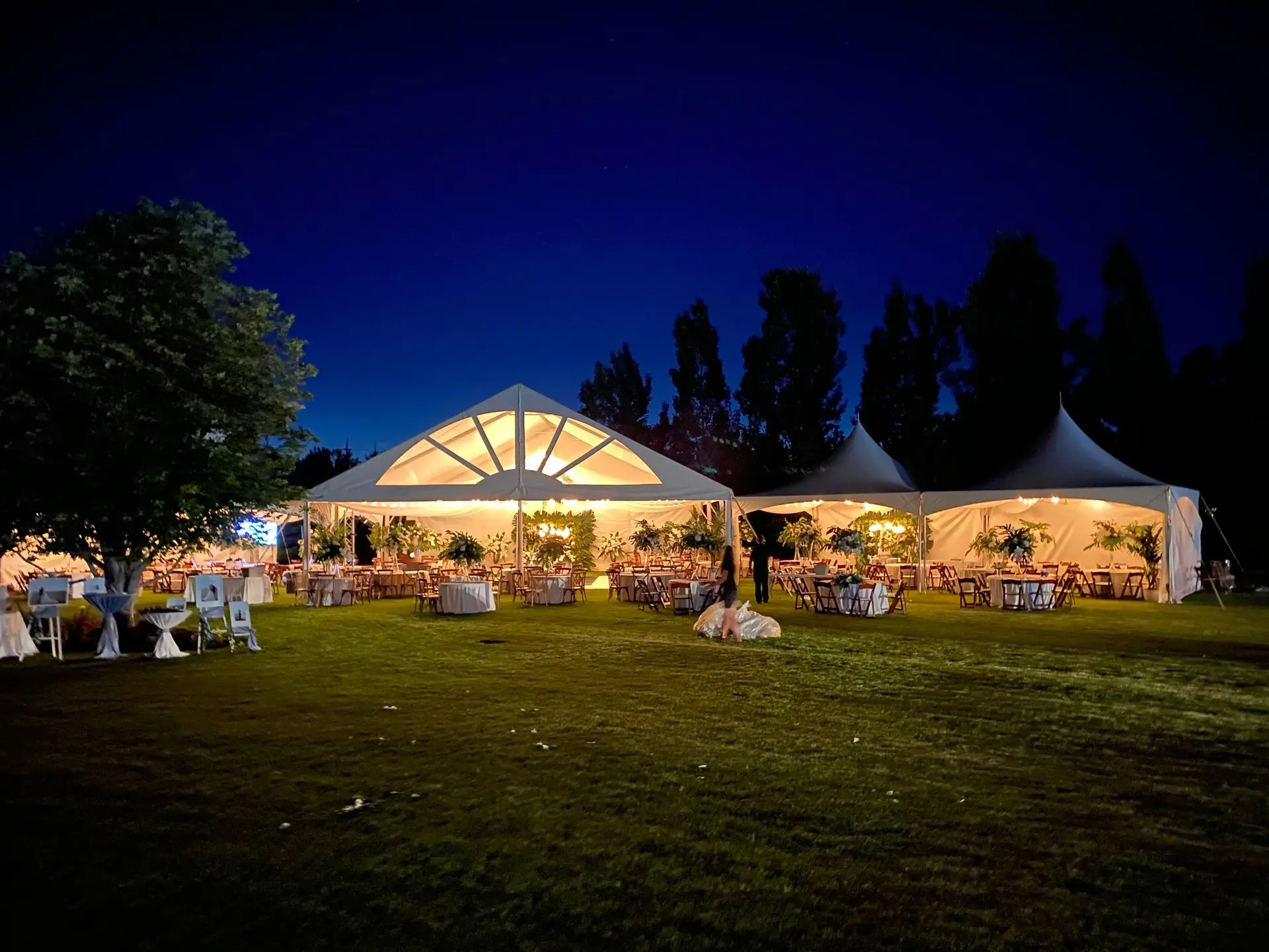 White marquee tents lit from inside at dusk on a wide lawn, set with tables for an evening reception