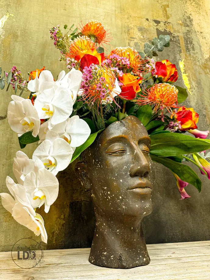 White orchids, orange proteas and red roses arranged in a bronze head sculpture on a wooden table