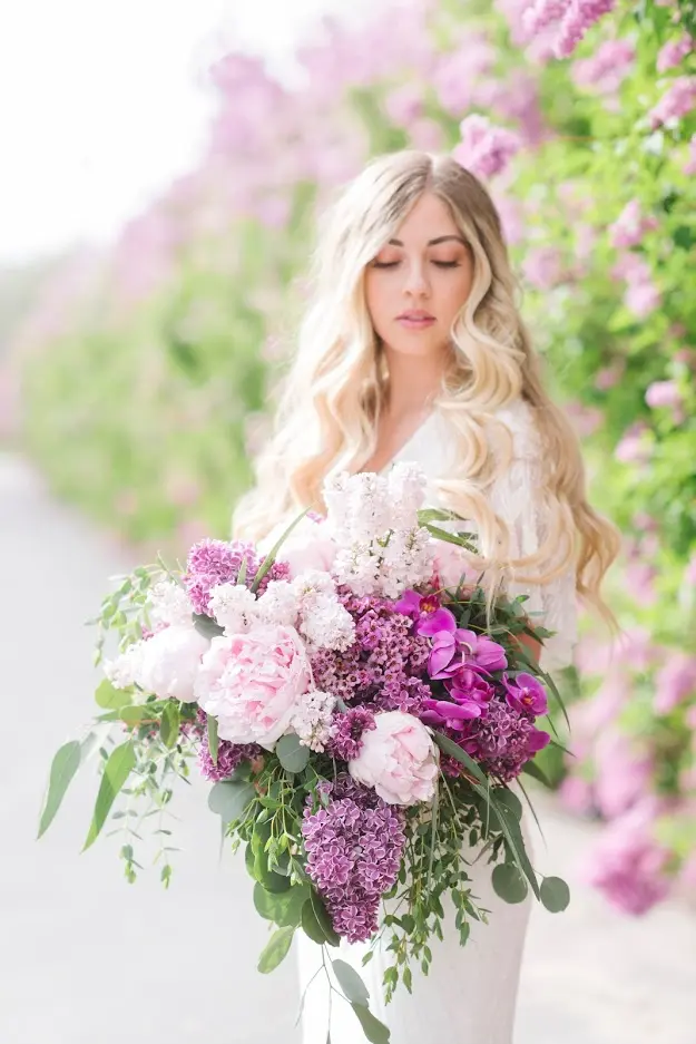 Bride holding a cascading bouquet of pink peonies, purple orchids and lilac along a path of flowering bushes